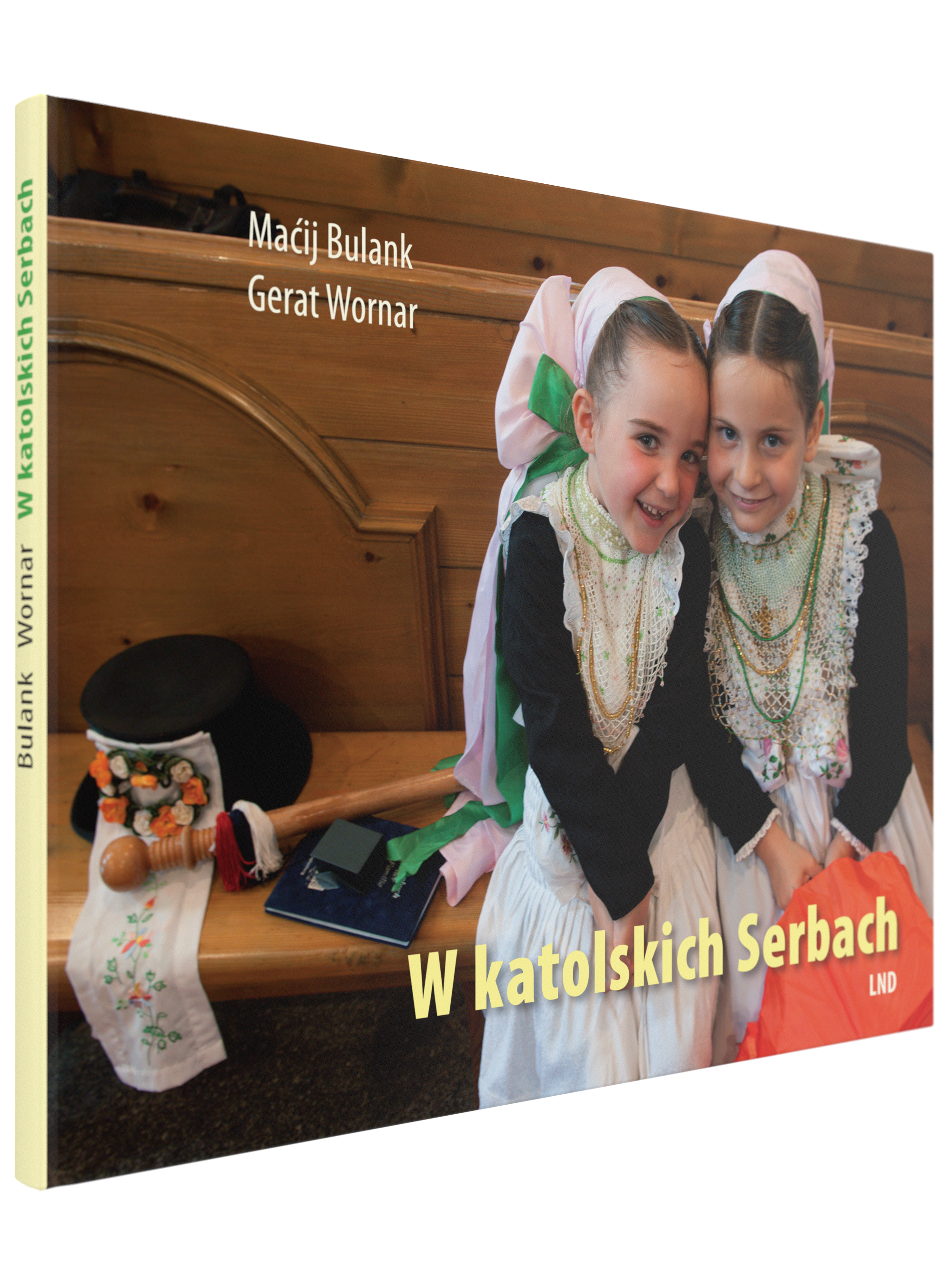 "Buchcover von 'W katolskich serbach' von Matthias Bulang und Gerhard Werner, erschienen im Domowina-Verlag. Das Cover zeigt zwei junge Mädchen in traditioneller sorbischer Tracht mit weißen Spitzenkragen, Perlenketten und großen Schleifen im Haar. Sie si